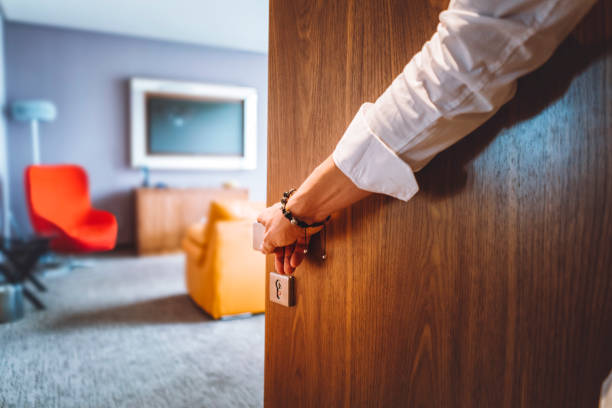 Toned man closing the door of the luxurious hotel room, man wearing a bracelet and a white shirt, closing the wooden door, nice and luxurious hotel room in the background.